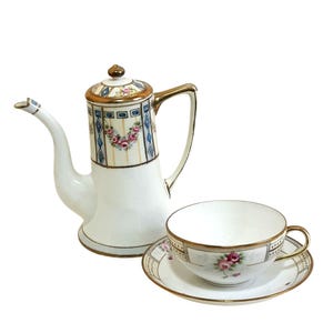 May include: A white porcelain tea set with gold trim, featuring a tall coffee pot and a teacup with saucer. The coffee pot has a decorative floral and geometric pattern in pink, blue, and gold. The teacup and saucer also have floral accents.