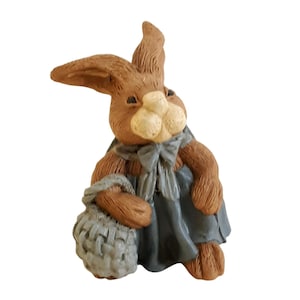 May include: A brown bunny figurine with a light-colored nose and a blue dress. The bunny is holding a gray basket. The figurine has a textured appearance and is likely made of ceramic or resin.