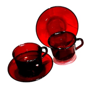 Può includere: Un set di tazze da tè e piattini in vetro rosso rubino. Il set comprende due tazze da tè con manici, due piattini e un piatto più grande. Il vetro è trasparente, permettendo alla luce di passare, ed è posto su uno sfondo bianco.