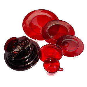 May include: A set of red glass dinnerware, including plates, saucers, and cups. The set features various sizes of plates and saucers, along with two cups. The glassware has a glossy finish and a deep red colour.