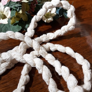 Pode incluir: Um close-up de uma corda de algodão branco, torcida e amarrada, com uma textura macia e natural. A corda está disposta em um padrão decorativo, destacando seu artesanato único.