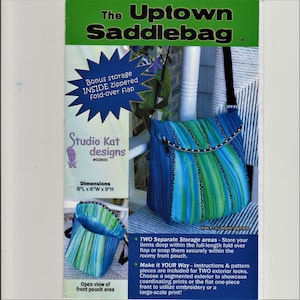May include: The Uptown Saddlebag pattern by Studio Kat Designs. The bag is blue and green with vertical stripes and a zippered fold-over flap. Dimensions are 20cm L x 15cm W x 23cm H.