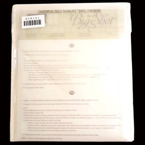 May include: A clear plastic package containing die-cutting templates. The package has the text "Cheerful Tags Thinlits Dies (141838)" and a barcode sticker. The Big Shot logo is visible. The package contains instructions and small circular elements.