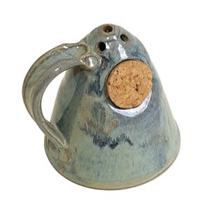 May include: A blue ceramic salt shaker with a cork top and a handle. The shaker has a speckled glaze and a rustic look.