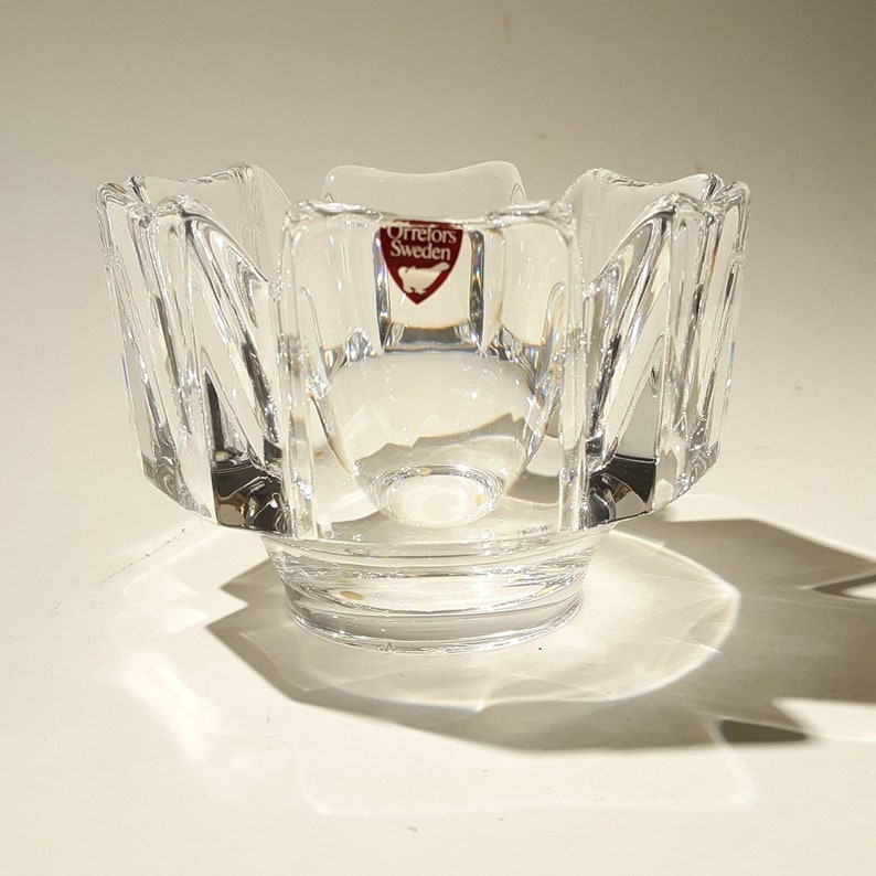 May include: A clear glass bowl with a scalloped edge and a starburst pattern. The bowl has a small, red label that reads "Orrefors Sweden".
