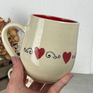 May include: Cream-colored ceramic mug with a red interior and a curved handle. The mug is decorated with three red hearts and black swirl designs. The mug is held in a hand.
