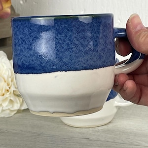 Könnte beinhalten: Handgefertigte Keramiktasse mit zweifarbigem Design. Die obere Hälfte ist ein gesprenkeltes, tiefes Blau, während die untere Hälfte weiß ist. Die Tasse hat eine abgerundete Form und einen stabilen Henkel, perfekt für Kaffee oder Tee.