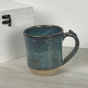 Può includere: Una tazza in ceramica fatta a mano con una glassa maculata turchese e marrone. La tazza ha una forma arrotondata con un manico robusto e una base naturale non smaltata. La tazza è alta circa 10 cm.