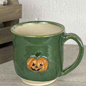 May include: A glossy forest green ceramic mug featuring a raised, three-dimensional orange jack-o'-lantern pumpkin design on the front. The mug has a smooth, rounded handle, a cream-colored interior, and a matching cream-colored base rim.