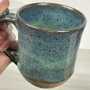 Może przedstawiać: Ręcznie wykonany ceramiczny kubek z nakrapianym niebiesko-zielonym szkliwem. Kubek ma brązową podstawę i uchwyt. Kubek jest trzymany przez dłoń. Unikalny element ceramiki, idealny do kawy lub herbaty.
