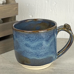 May include: A ceramic mug featuring a deep blue and brown reactive glaze finish. The upper portion displays a mottled blue texture, transitioning into a darker brown base. The mug has a sturdy handle and a plain, unglazed cream-colored bottom rim.