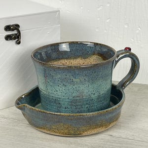 Puede incluir: Un juego de taza y platillo de cerámica hecho a mano. La taza y el platillo están esmaltados en tonos azules y marrones. La taza tiene un asa con un pequeño acento rojo. El platillo tiene un borde curvo. Para servir té o café.