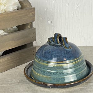 May include: A ceramic butter dish featuring a dome-shaped lid with a looped handle. The piece has a glossy glaze in shades of deep blue and teal, with horizontal ridges circling the lid. The base plate has a dark brown rim, matching the handle.