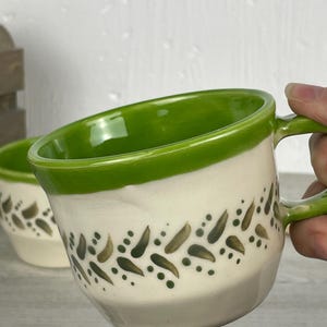 May include: A ceramic mug with a cream-colored base and a bright green rim. The exterior features a hand-painted pattern of dark green leaf-like shapes and small dots encircling the middle. The interior of the mug is glazed in a vibrant green.