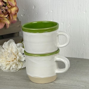 May include: Two white ceramic mugs stacked on top of each other. Each mug has a green rim and a beige base. The mugs have a simple, handmade appearance with a curved handle.