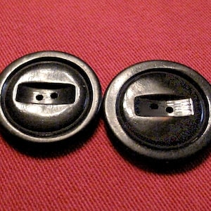 May include: Two dark gray, round buttons with a rectangular center and two small holes. The buttons have a slightly raised edge and are set against a red background.
