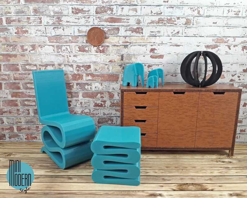 Puede incluir: Un conjunto de silla y otomana moderna en miniatura de color azul turquesa. La silla tiene un dise&ntilde;o &uacute;nico y ondulado. La otomana est&aacute; apilada con tres capas. El conjunto se muestra frente a una c&oacute;moda en miniatura con cajones y una escultura de esfera negra.