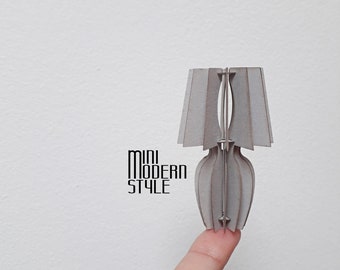 May include: A miniature gray cardboard lamp with a pleated shade and a cylindrical base. The lamp is held by a finger.
