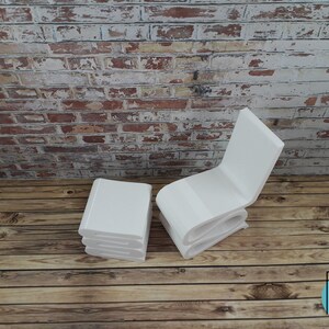 Puede incluir: Conjunto de silla y otomana en miniatura de color blanco, hechos con formas de libros apilados. La silla y la otomana est&aacute;n sobre un suelo de madera con una pared de ladrillo en el fondo.