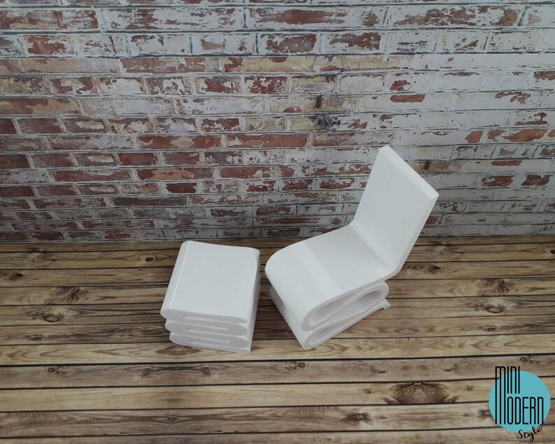 Puede incluir: Conjunto de silla y otomana en miniatura de color blanco, hechos con formas de libros apilados. La silla y la otomana est&aacute;n sobre un suelo de madera con una pared de ladrillo en el fondo.