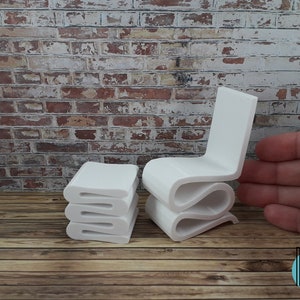 Puede incluir: Conjunto de silla y otomana en miniatura de color blanco. La silla y la otomana est&aacute;n hechas de un material blanco liso y tienen un dise&ntilde;o moderno y abstracto. La silla tiene un respaldo y un asiento curvos, y la otomana es una pila de tres piezas rectangulares.