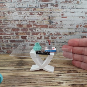 May include: A white modern coffee table with a clear glass top. The table has a unique X-shaped base and is decorated with a teal lampshade, a small gray vase with red sticks, and a book. The table is approximately 15 inches long.