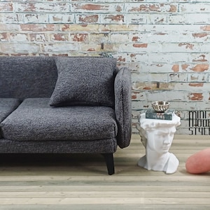 Puede incluir: Un sofá gris de tela con un cojín se encuentra frente a una pared de ladrillo blanco. Un pequeño busto blanco con un libro y un tazón en la parte superior está en primer plano. El texto "Mini Modern Style" es visible en la imagen.