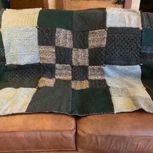 Pode incluir: Uma colcha de retalhos com vários tecidos de malha texturizados em tons de verde, cinza e marrom. A colcha é adornada com botões de madeira e está drapeada sobre um sofá de couro marrom. O design apresenta quadrados e retângulos de diferentes padrões de malha.