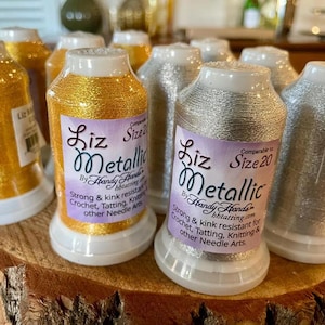 May include: A collection of Liz Metallic thread spools in gold and silver colors. Each spool features a white label with the text Liz Metallic by Handy Hands, Size 20, and descriptions for crochet, tatting, and knitting applications.