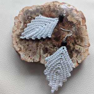 May include: A pair of white crocheted earrings with black lava rock beads hanging from silver hooks.