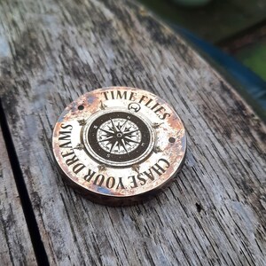 Könnte beinhalten: Ein runder, kupferfarbener Metallanhänger mit einem Kompass-Design und dem Text "CHASE YOUR DREAMS TIME FLIES", der um den Rand graviert ist.