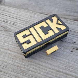 May include: A rectangular black and gold object featuring the word SICK in bold, raised lettering on the top surface. The item has a layered, brick-like texture on the sides and includes a small, protruding gold tab extending from the front edge.