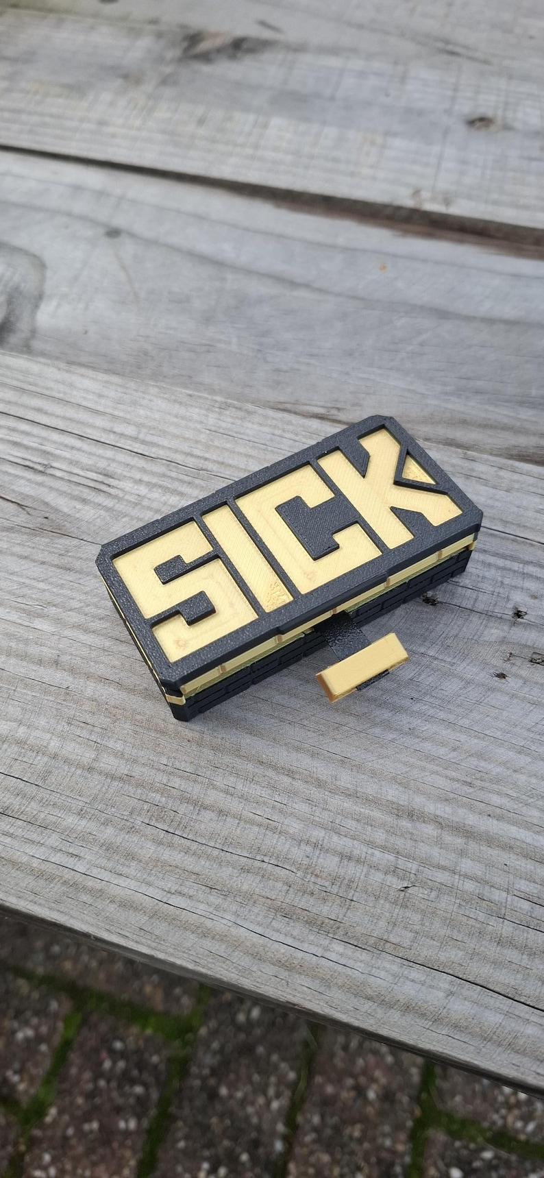 May include: A rectangular black and gold object featuring the word SICK in bold, raised lettering on the top surface. The item has a layered, brick-like texture on the sides and includes a small, protruding gold tab extending from the front edge.