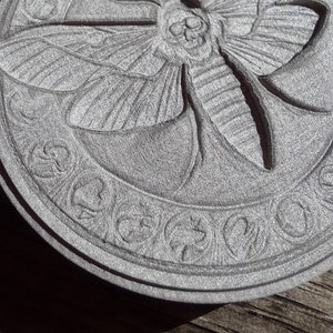 May include: A circular, gray, stone coaster with a detailed, carved design of a moth with a skull on its head. The moth is surrounded by a decorative border.