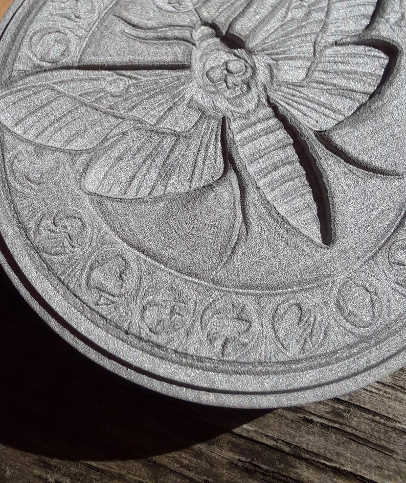 May include: A circular, gray, stone coaster with a detailed, carved design of a moth with a skull on its head. The moth is surrounded by a decorative border.