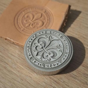 Könnte beinhalten: Ein silberner Metallstempel mit einem Fleur-de-Lis-Design, neben einem Stück gestempeltem Leder. Der Stempel ist rund mit einem detaillierten Rand. Das Leder zeigt einen geprägten Abdruck des gleichen Designs.