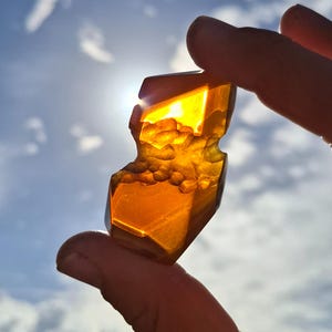 Puede incluir: Una piedra preciosa translúcida de color ámbar sostenida entre los dedos contra un cielo brillante. La piedra tiene una superficie rugosa y texturizada con un borde pulido y facetado. La luz del sol brilla a través de la piedra, creando un efecto brillante.