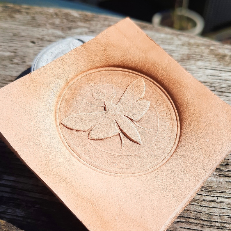 May include: A close-up of a light brown leather stamp with a detailed embossed design of a moth with intricate wings. The stamp is circular and has a decorative border.
