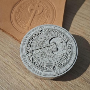 Puede incluir: Un sello metálico circular con un diseño detallado de hacha, rodeado por un borde decorativo. El sello está junto a un trozo de cuero estampado, que muestra la impresión del diseño. El sello está sobre una superficie de madera.