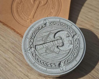 May include: A circular metal stamp with a detailed axe design, surrounded by a decorative border. The stamp is next to a piece of stamped leather, showing the impression of the design. The stamp is on a wooden surface.