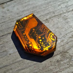Può includere: Un oggetto esagonale unico con un design a circuito stampato. L'oggetto presenta una combinazione di colori nero e arancione, con motivi e dettagli intricati che ricordano i componenti elettronici. È posto su una superficie di legno invecchiato.
