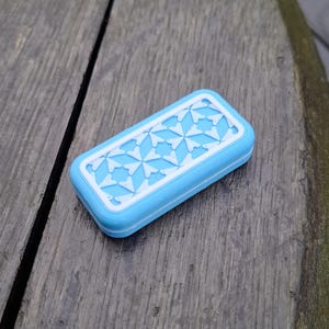 May include: A rectangular, light blue soap dish with a white, geometric starburst pattern on the lid. The dish is made of a smooth material and sits on a weathered wooden surface.