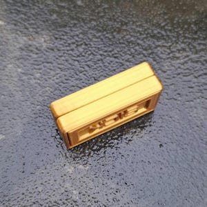 May include: A rectangular, gold-coloured object with a smooth top surface and a recessed side panel featuring embossed, stylised lettering. The item has a layered, block-like construction with visible horizontal lines along its edges and sides.