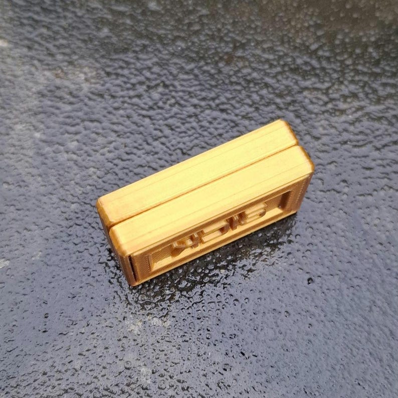 May include: A rectangular, gold-coloured object with a smooth top surface and a recessed side panel featuring embossed, stylised lettering. The item has a layered, block-like construction with visible horizontal lines along its edges and sides.