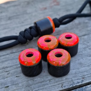 Puede incluir: Un conjunto de cuentas cilíndricas negras con tapas rosas y naranjas vibrantes, con un agujero central. Un cordón negro a juego está unido a una cuenta más grande con detalles en naranja. Las cuentas están dispuestas sobre una superficie de madera envejecida.