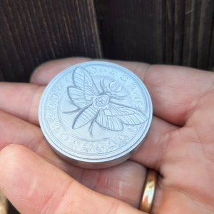 May include: A silver-colored metal coin with a detailed engraving of a moth with a skull on its head. The moth is surrounded by a circle of symbols and text.