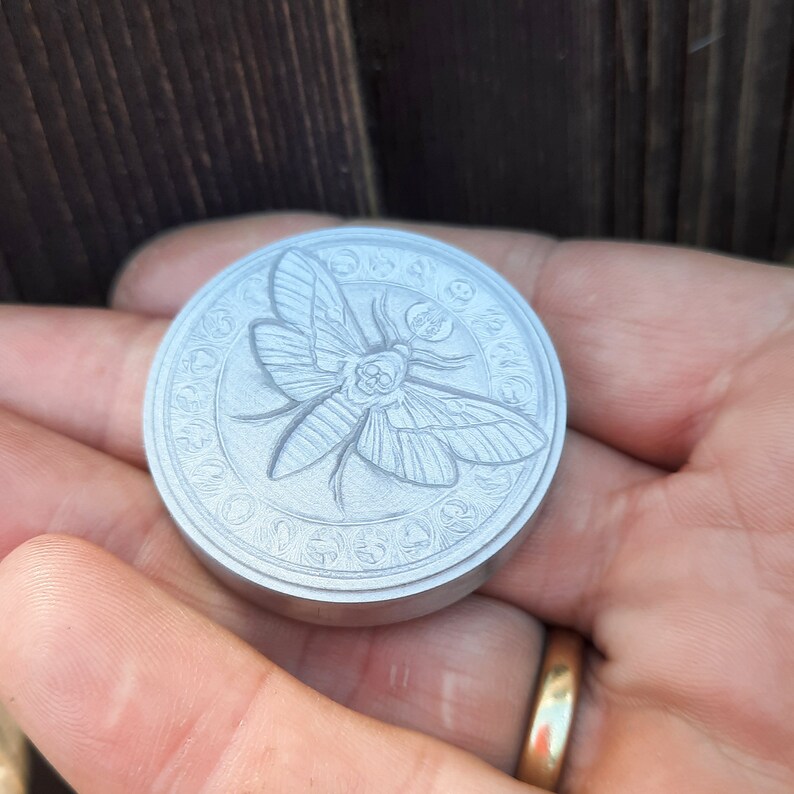 May include: A silver-colored metal coin with a detailed engraving of a moth with a skull on its head. The moth is surrounded by a circle of symbols and text.