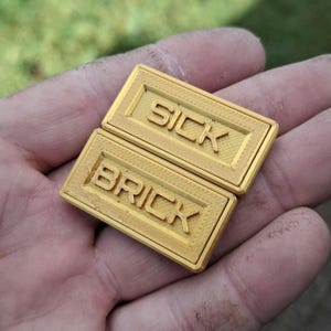 May include: Two rectangular gold-coloured plastic bricks with a textured surface. The top brick features the embossed text SICK, and the bottom brick features the embossed text BRICK. Both items have a raised border frame around the lettering.