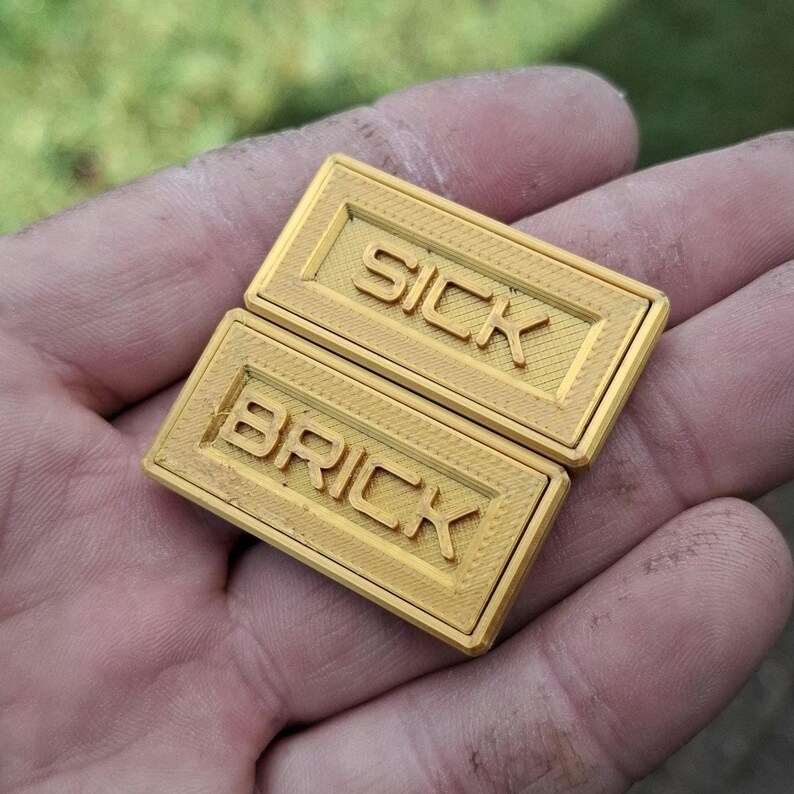 May include: Two rectangular gold-coloured plastic bricks with a textured surface. The top brick features the embossed text SICK, and the bottom brick features the embossed text BRICK. Both items have a raised border frame around the lettering.