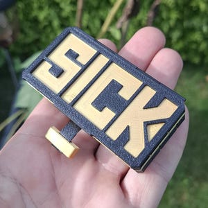 May include: A rectangular 3D printed sign featuring the word SICK in raised gold lettering against a textured black background. The object includes a small gold and black attachment extending from the bottom edge, showcasing a layered construction.